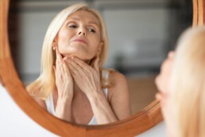 Middle-aged woman examining her neck in the mirror at home, gently touching her skin after double chin treatment or neck lift for a firmer, more youthful appearance.