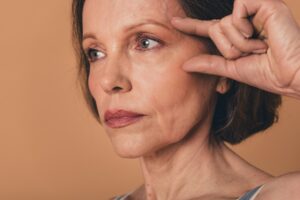 Close-up photo of a senior woman with no filter, gently touching her face and contemplating a facelift, isolated on a beige background.