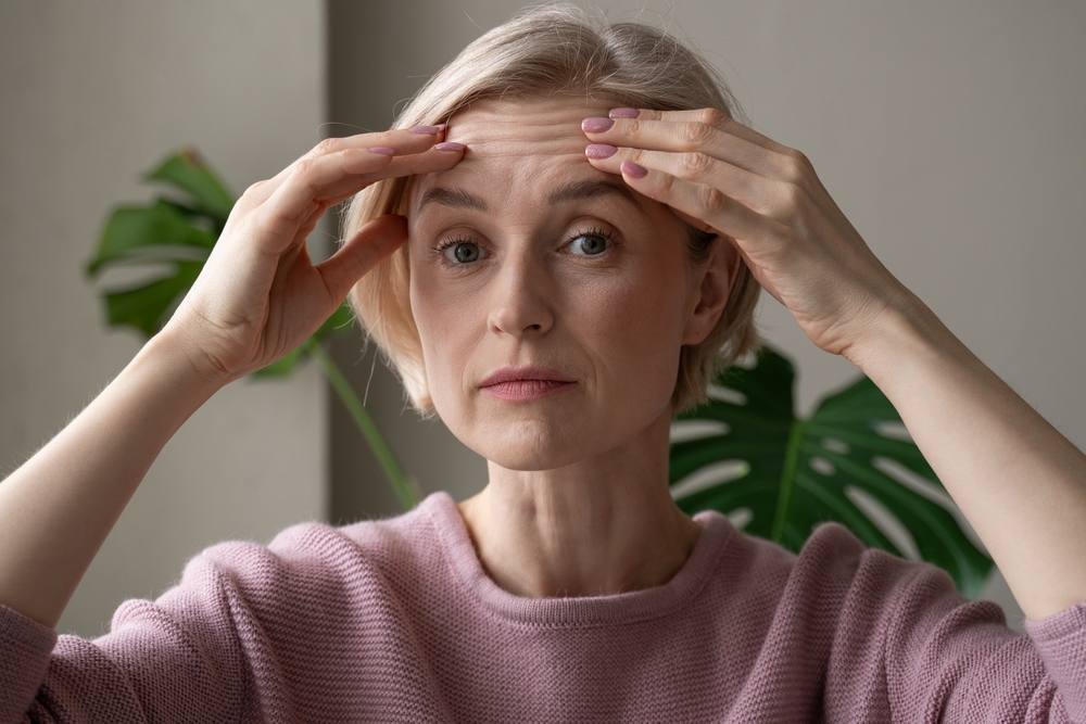 Contemplative mature woman indoors gently touching her forehead with both hands, expressing concern or introspection, illustrating the concept of a brow lift.