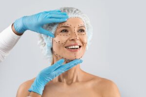 Cheerful middle-aged woman with pre-surgery face marks wearing medical hat, smiling as surgeon in blue gloves touches her skin before cosmetic procedure.