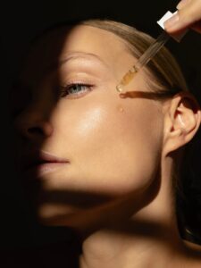 Close-up of woman’s glowing face with glass pipette applying luminous cosmetic serum, concept of luxury skincare, hydration, and beauty self-care