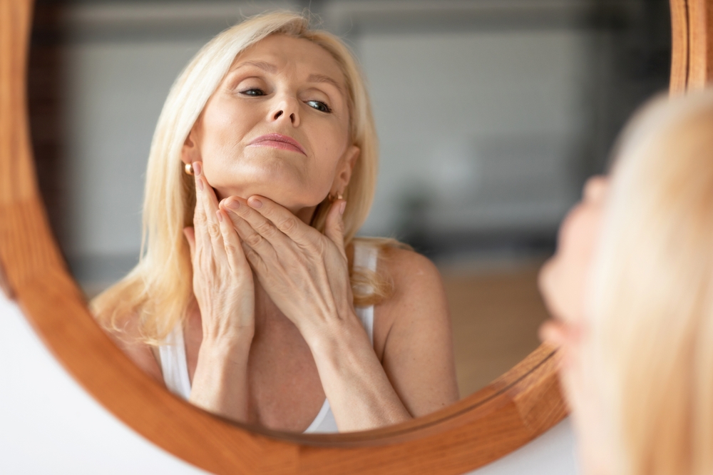 1770823603_shutterstock_2353711721 Middle-aged woman touching the smooth skin on her neck while looking in the mirror at home, highlighting double chin treatment and neck lift results.